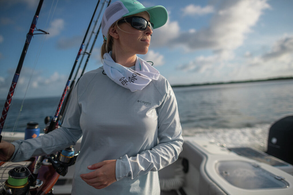 Mulher em barco de pesca usando roupas Columbia com proteção UV e protetor de pescoço, exemplo de como montar uma mala de pescaria com itens essenciais.