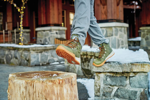 Pessoa caminhando em ambiente urbano nevado usando uma das melhores botas para neve Columbia, modelo verde com solado antiderrapante laranja.
