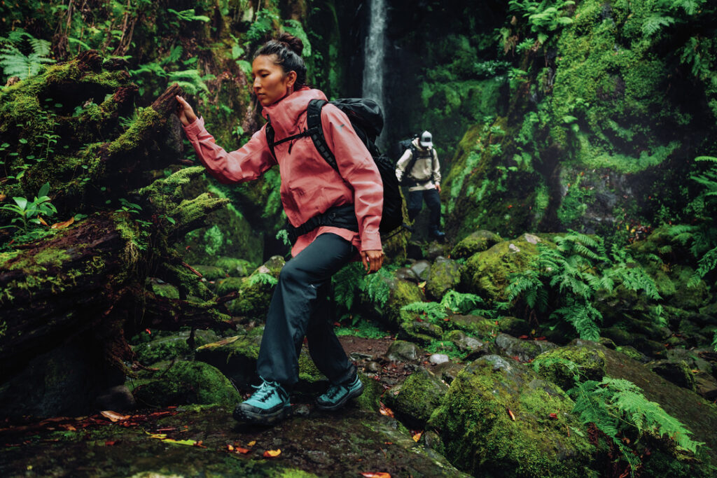 Mulher usando jaqueta impermeável Columbia rosa em trilha na floresta úmida, com mochila de trekking, praticando hiking sob chuva leve.
