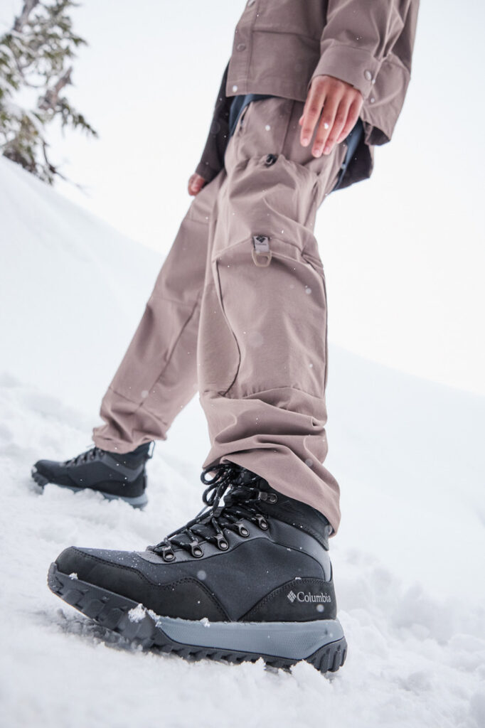 Homem com bota Columbia masculina caminhando em floresta com neve caindo.