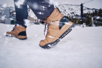 Homem usando bota Columbia masculina em ambiente de neve com sol e montanhas ao fundo.