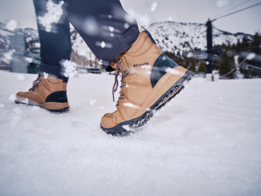 Homem usando bota Columbia masculina em ambiente de neve com sol e montanhas ao fundo.
