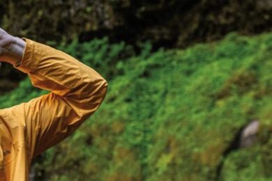 Homem usando jaqueta impermeável amarela Columbia em trilha chuvosa, com vegetação verde ao fundo; ao fundo, mulher com jaqueta vermelha impermeável.