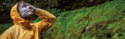 Homem usando jaqueta impermeável amarela Columbia em trilha chuvosa, com vegetação verde ao fundo; ao fundo, mulher com jaqueta vermelha impermeável.