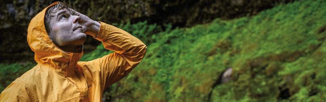 Homem usando jaqueta impermeável amarela Columbia em trilha chuvosa, com vegetação verde ao fundo; ao fundo, mulher com jaqueta vermelha impermeável.