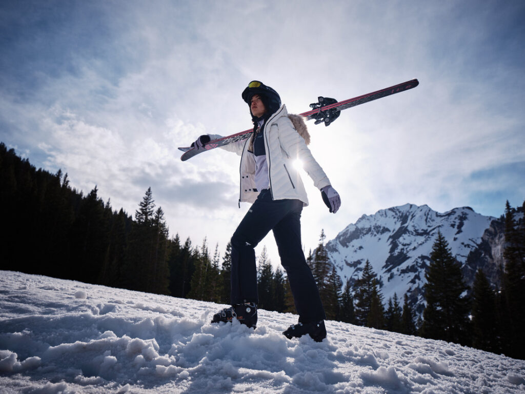 Pessoa usando roupa de ski Columbia branca com esquis nos ombros em cenário de montanha nevada.